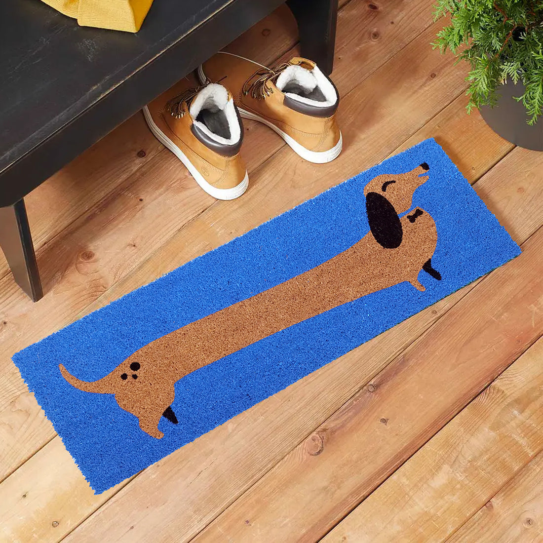 A blue doormat with a brown and black dachshund design is placed on a wooden floor next to a pair of sneakers.