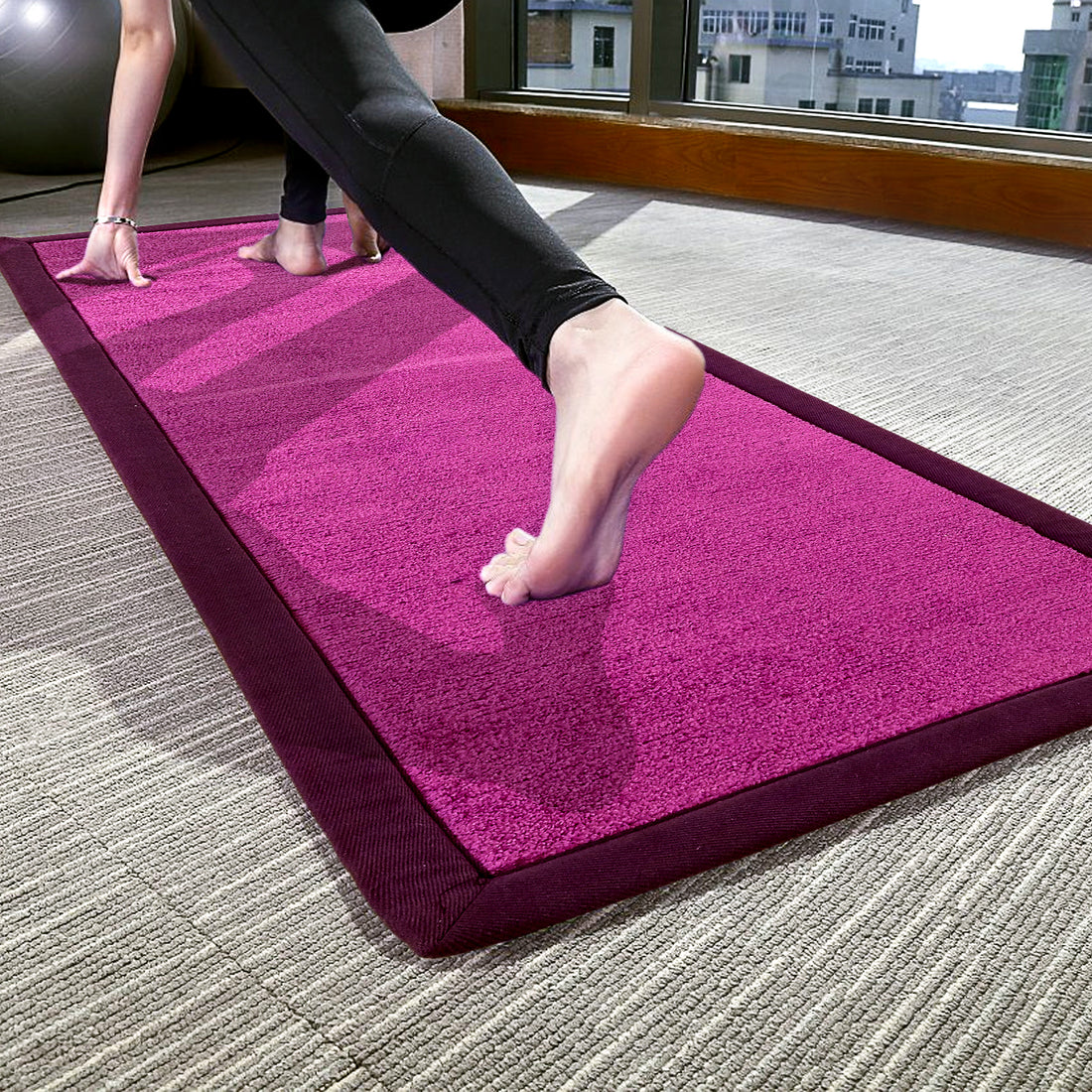 A person practising yoga on a long purple-colored oblong yoga mat with a cotton border, placed on a carpeted floor.