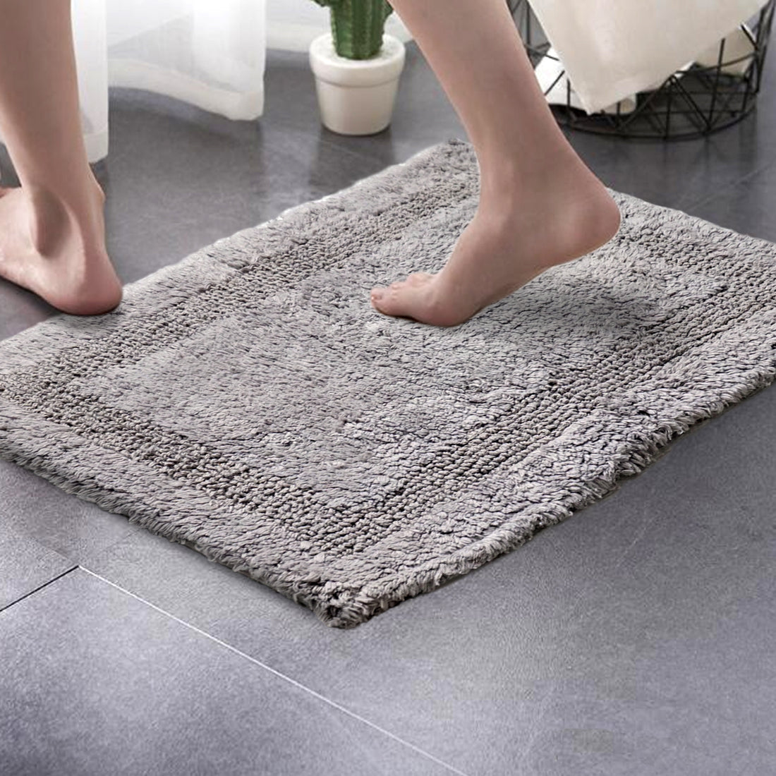 A person's feet stepping onto a grey, solid-patterned cotton bath mat placed on a tiled floor.