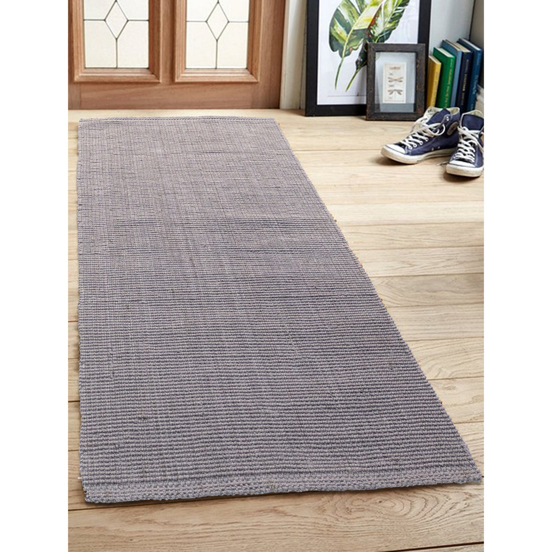 A light grey, flatweave jute runner rug is placed on a wooden floor, with a pair of sneakers at the top of the image indicating the scale of the rug.