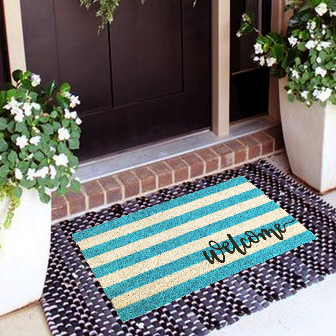 A rectangular natural coir door mat with a blue and yellow striped pattern and the word 'WELCOME' printed on it, placed on a doorstep with a brick wall and potted plants in the background.