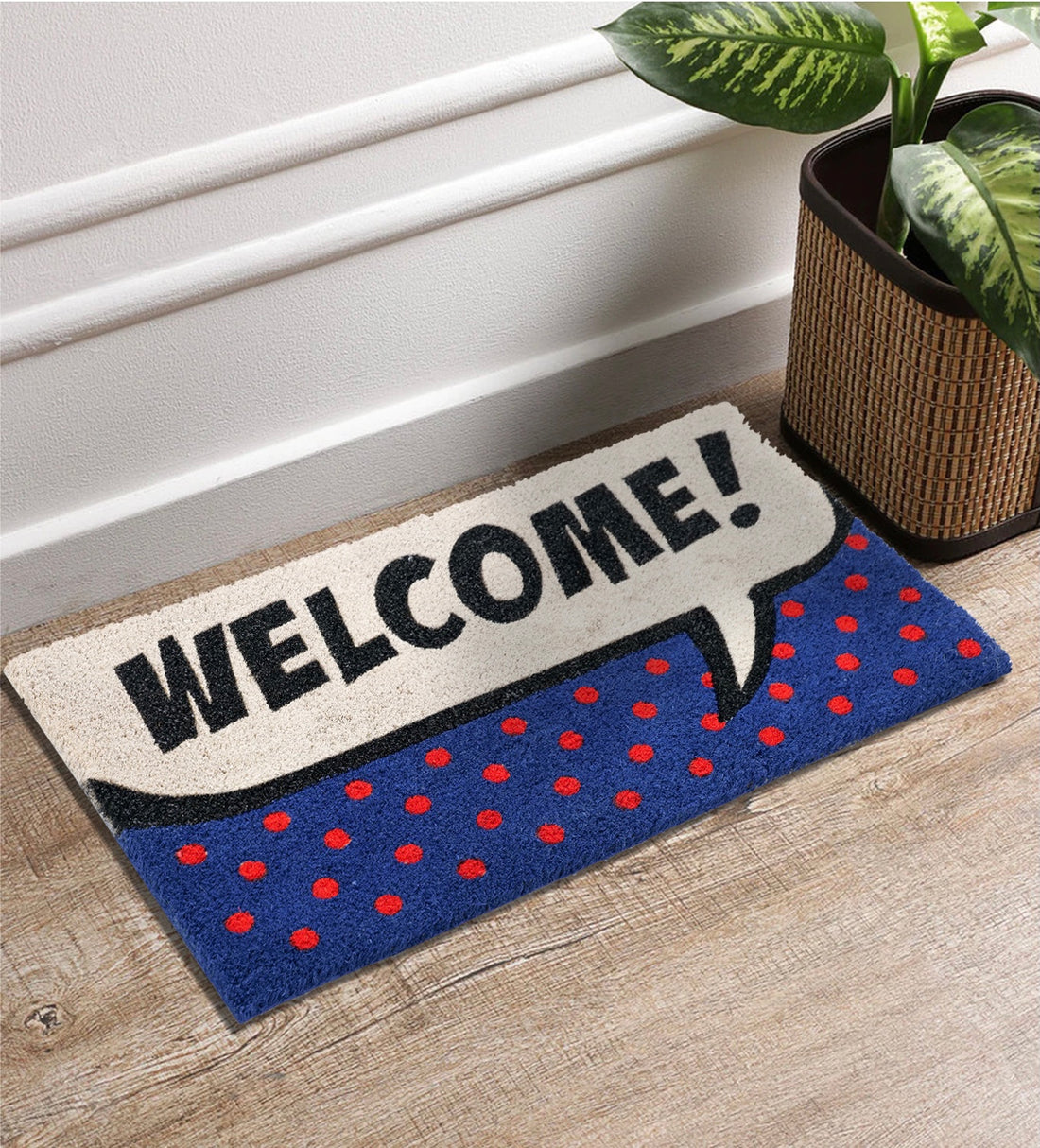 Rectangular natural coir door mat with a pop art fun design featuring the text 'Welcome!' in a speech bubble style, decorated with blue and red polka dots, placed on a wooden floor by a plant pot.