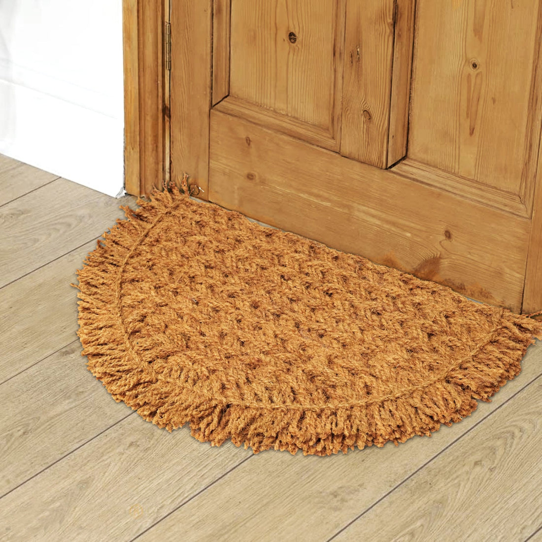A semi-circle shaped, handwoven coir rope doormat with fringed edges, placed on a wooden floor in front of a door.