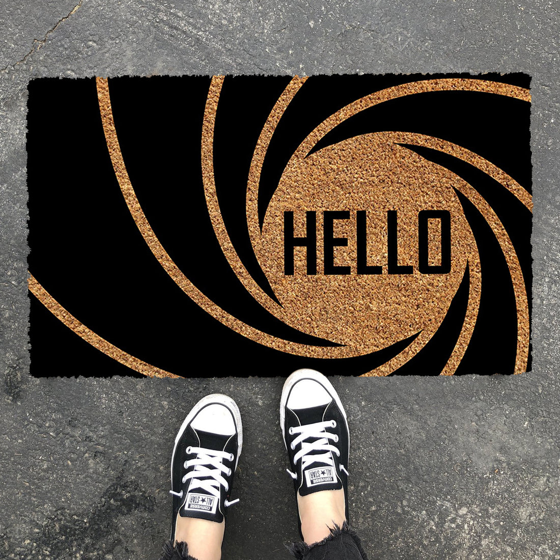 A rectangular natural coir doormat with 'Hello' text and a 007 bond theme design, placed on the ground with a pair of sneakers at the top of the image.