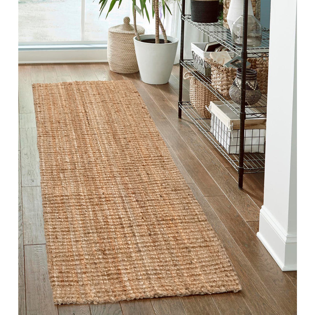 A natural handspun jute runner rug in a plain weave design, placed on a wooden floor beside a metal and wicker shelf, with a potted plant and various decorative items in the background.