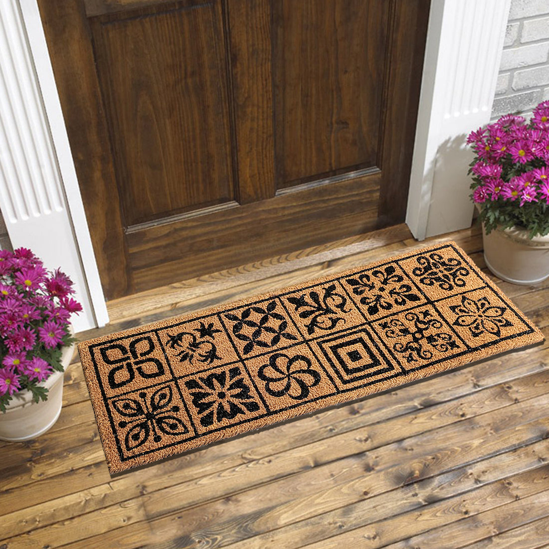 A rectangular natural coir doormat with a geometric mosaic tile pattern, placed on a wooden floor in front of a door, with potted flowers on either side.