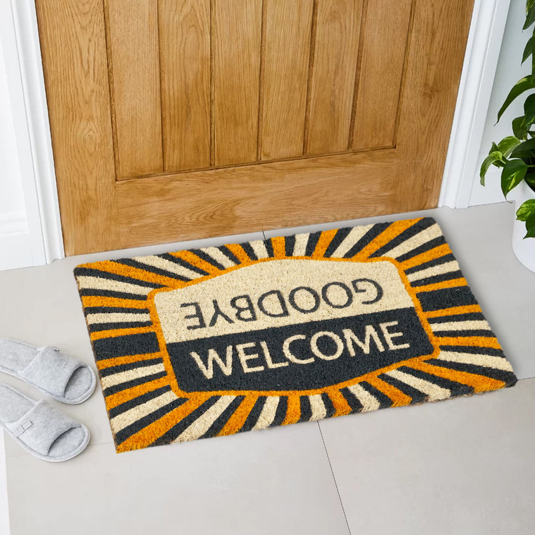 A rectangular natural coir doormat with a yellow and grey geometric pattern and the text 'GOOD BYE' and 'WELCOME' on it. The mat is placed on the floor with a pair of slippers at its bottom left corner.