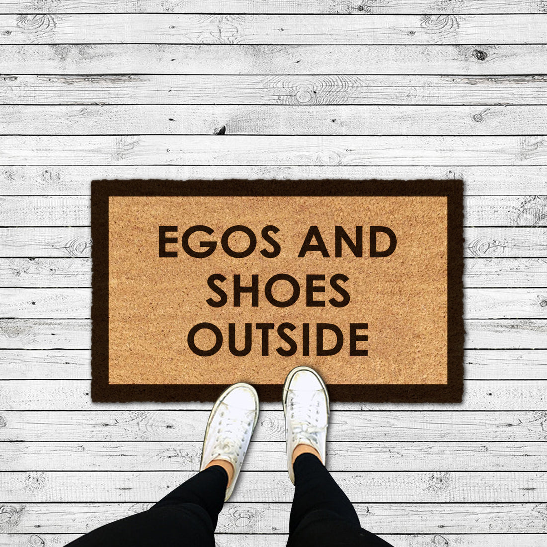 A rectangular natural coir doormat with the text 'EGOS AND SHOES OUTSIDE' printed in bold capital letters, placed on a wooden floor with a person's feet in white shoes at the bottom edge of the image.