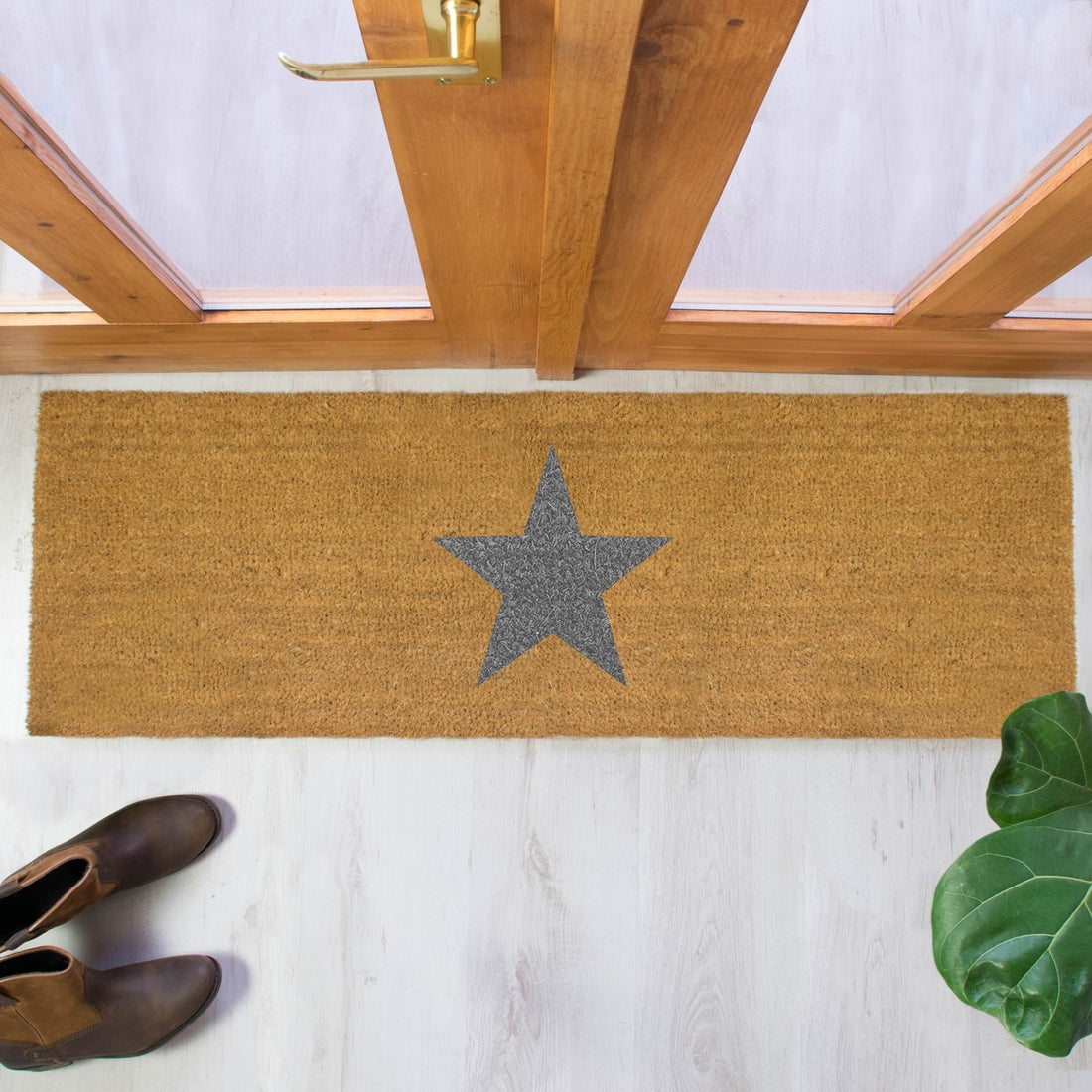 A rectangular natural coir doormat with a gray star printed design, placed on a wooden floor, with a pair of boots at the bottom of the image and a plant to the side.
