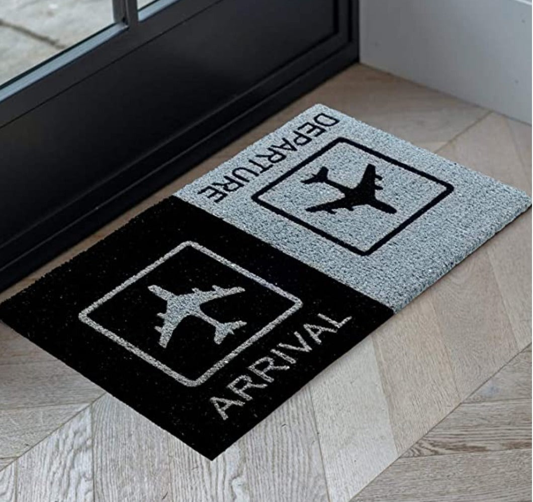 A rectangular natural coir doormat with black and grey geometric patterns, featuring an aeroplane symbol and the words 'ARRIVAL'  and 'DEPARTURE' on a dark floor, placed on a doorstep.