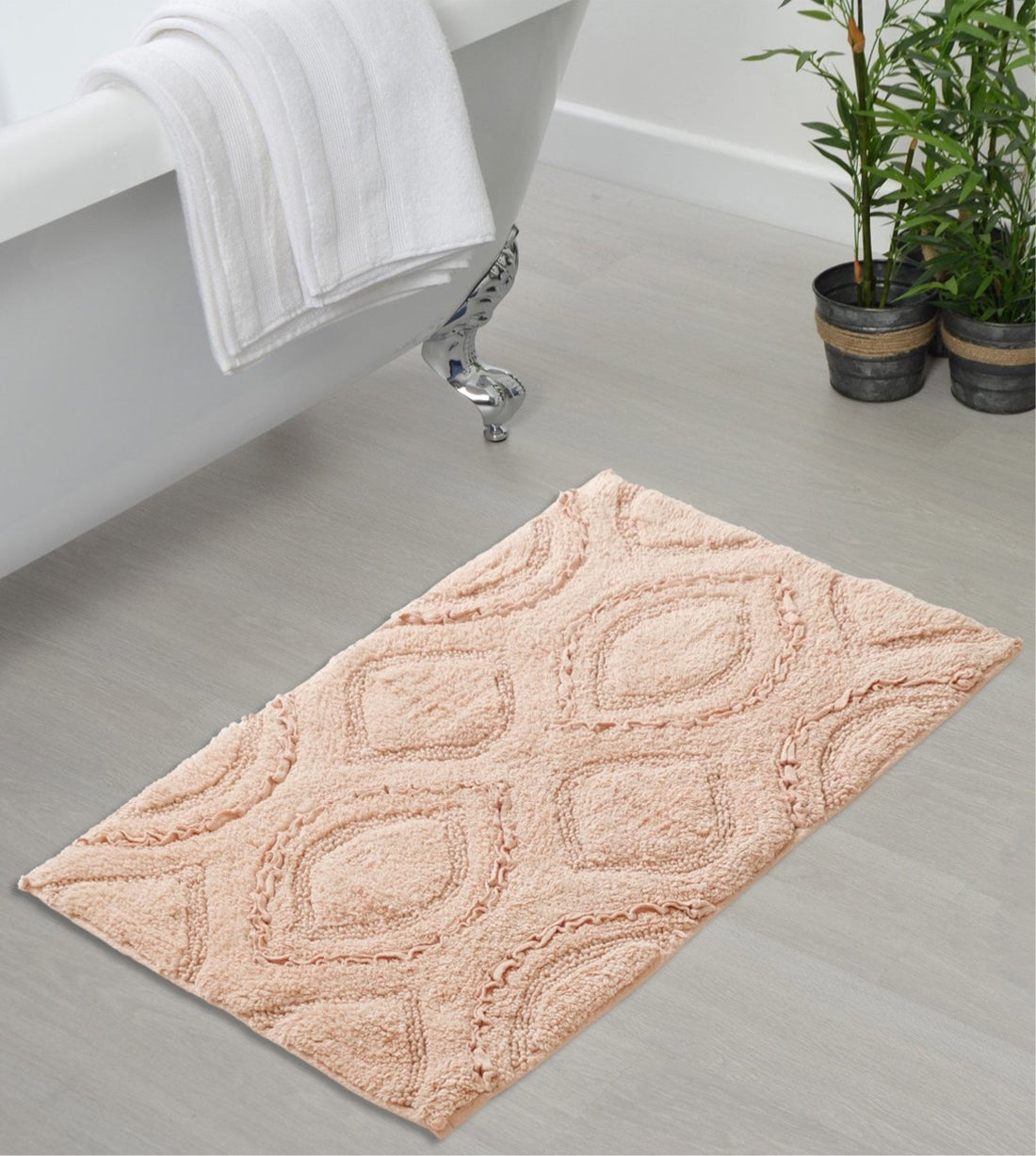 A woven cotton bath mat in peach colour, with a geometric pattern, is placed on a bathroom floor near a bathtub and a potted plant.