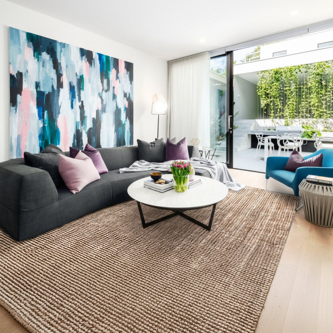 
                      
                        A modern living room featuring a grey sofa with purple cushions, a white round side table with a lamp, a woven jute rug, and a large abstract painting on the wall.
                      
                    