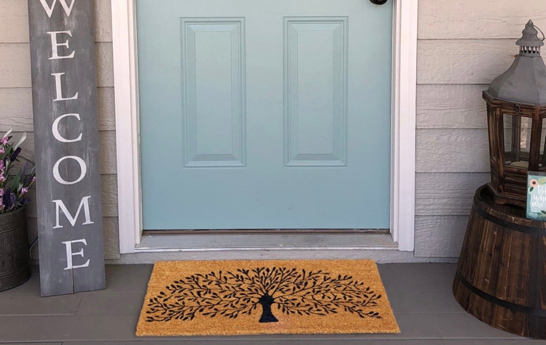 
                      
                        A rectangular natural coir doormat with a printed design featuring trees, placed on the ground in front of a blue door, with a gray sign that reads 'Welcome' and a decorative bottle on the side.
                      
                    
