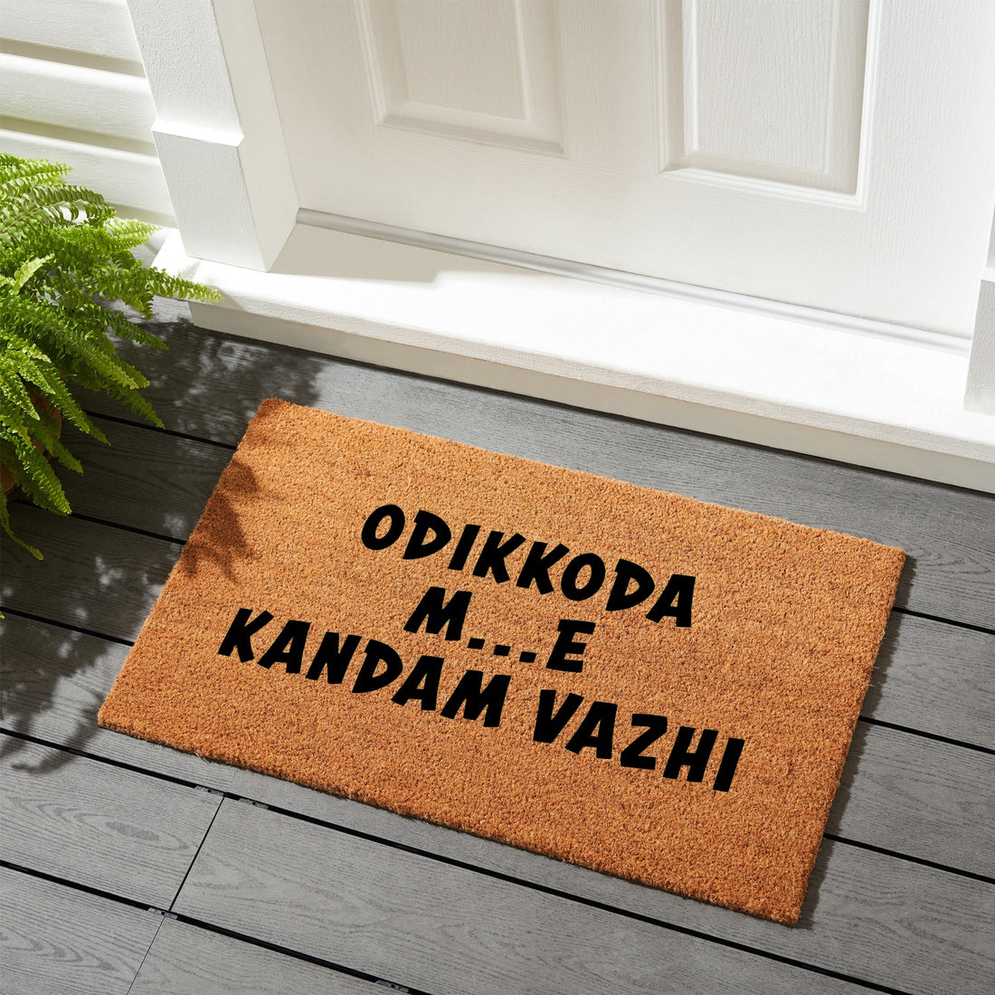 A rectangular natural coir door mat with a black printed design in Malayalam script, placed on a wooden floor, with a potted plant to the left and a white door in the background.