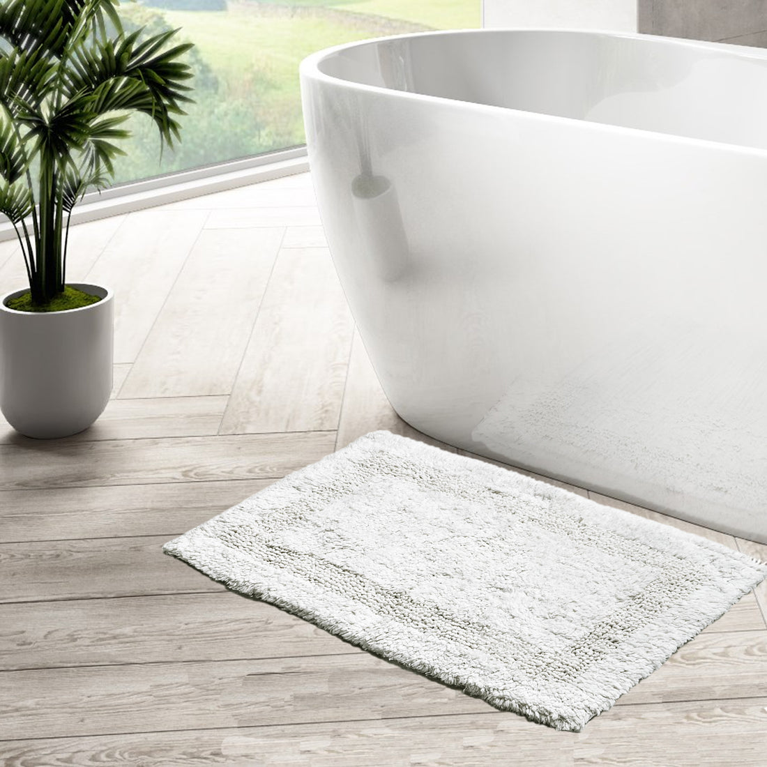 A white cotton bath mat with a textured surface, placed on a tiled bathroom floor in front of a bathtub, with a potted plant and wooden flooring visible in the background.