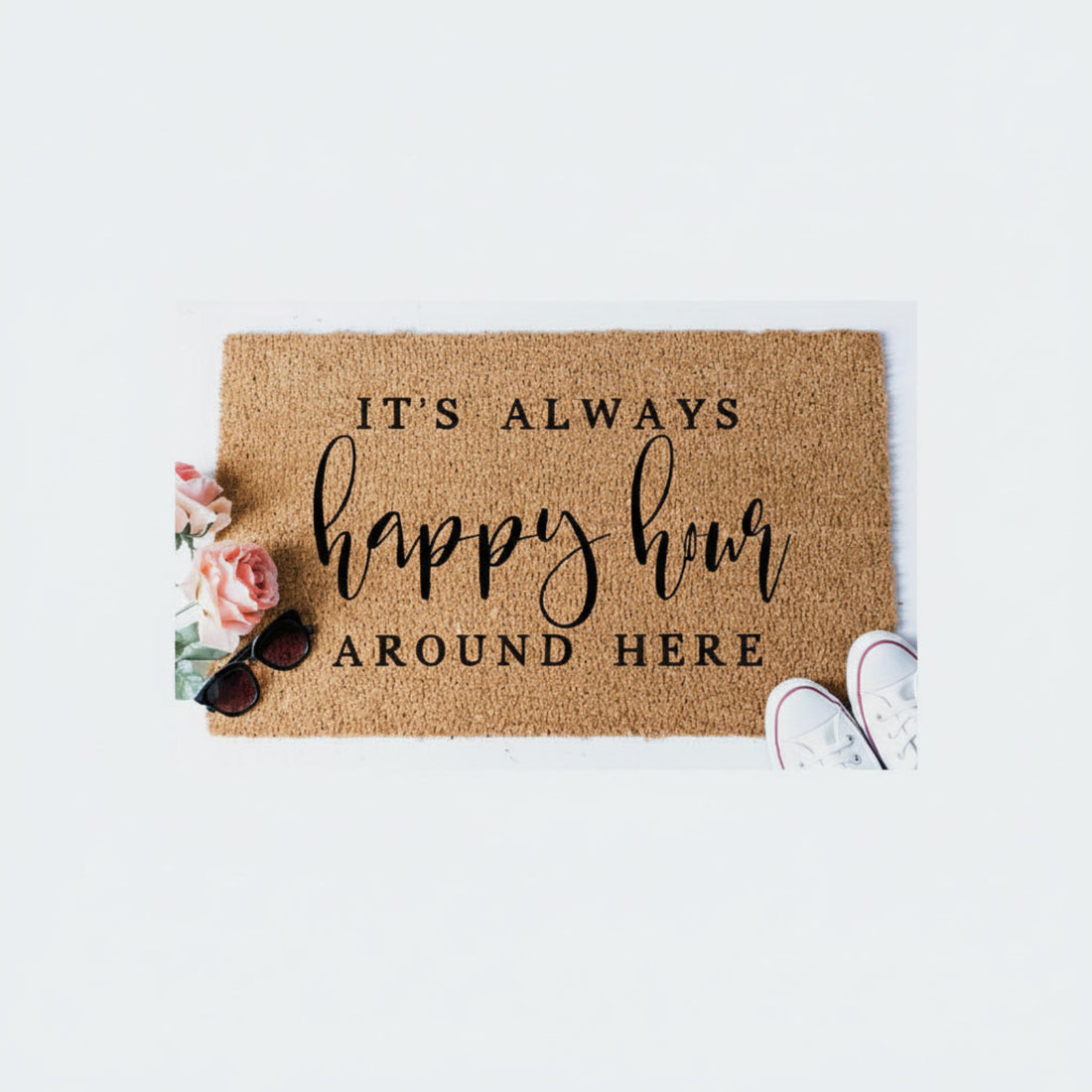 Rectangular natural coir floor mat with a printed design that reads 'It's always happy hour around here' in cursive script, placed on a white surface with a pair of sunglasses and a shoe on top of it.