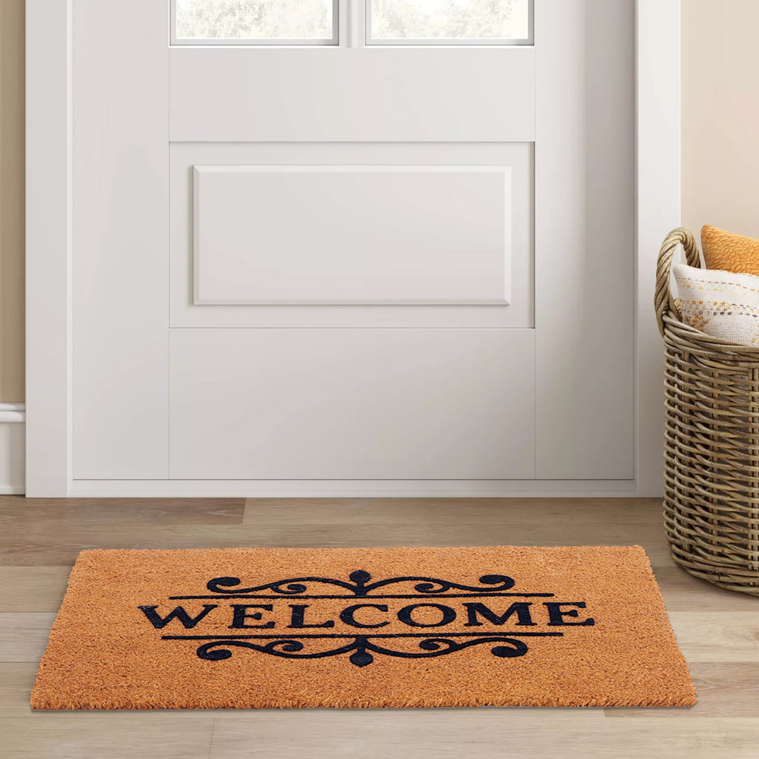 A rectangular coir doormat with 'Welcome' text flocked on it, placed on a wooden floor in front of a white door, with a wicker basket in the background.