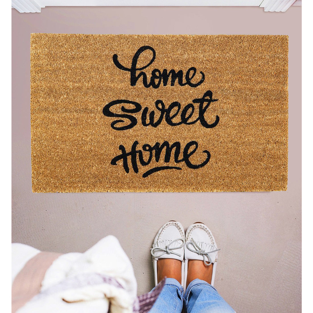 A rectangular natural coir door mat with the phrase 'HOME SWEET HOME' printed in black. The mat is placed on the floor, and there is a person standing on it wearing casual shoes.