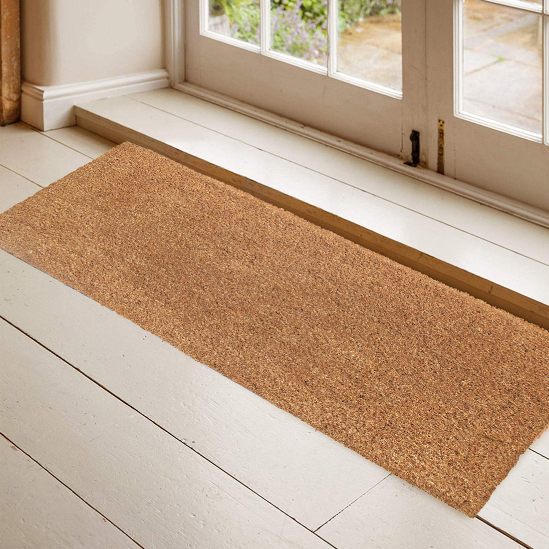 A rectangular natural plain coir door mat with anti-slip backing is placed on a white floor by the entrance of a house.