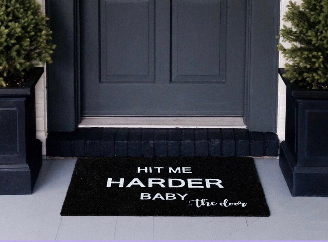 Black doormat with 'Hit Me Harder Baby' text in front of a dark gray door.