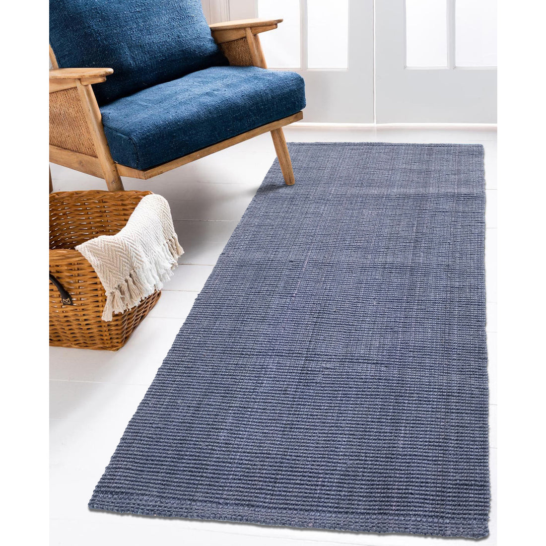 A dark grey, flatweave jute runner rug laid out on a white floor next to a blue couch with a person sitting on it, with a woven basket and a shawl in the background.
