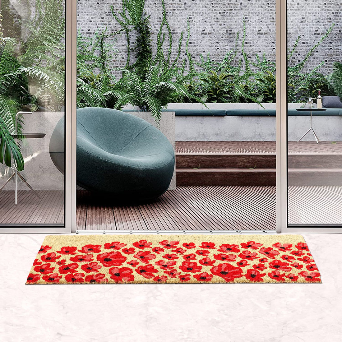 A rectangular, flower-printed natural coir entrance door mat placed on a floor, with a large glass door leading to a decked area with plants and a modern chair.