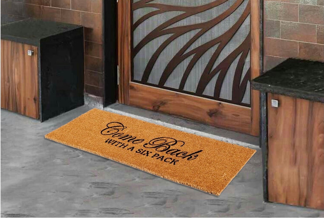 A large natural coir oblong doormat with a humorous message 'Come back with a six pack' printed on it, placed on the floor in front of a wooden door and a black bench, with a zebra-stripes pattern on the door in the background.