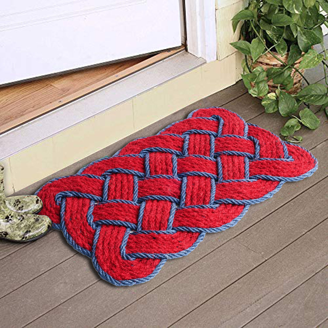 A red and blue geometric patterned coir rope floor mat placed on a wooden floor with a green plant beside it.