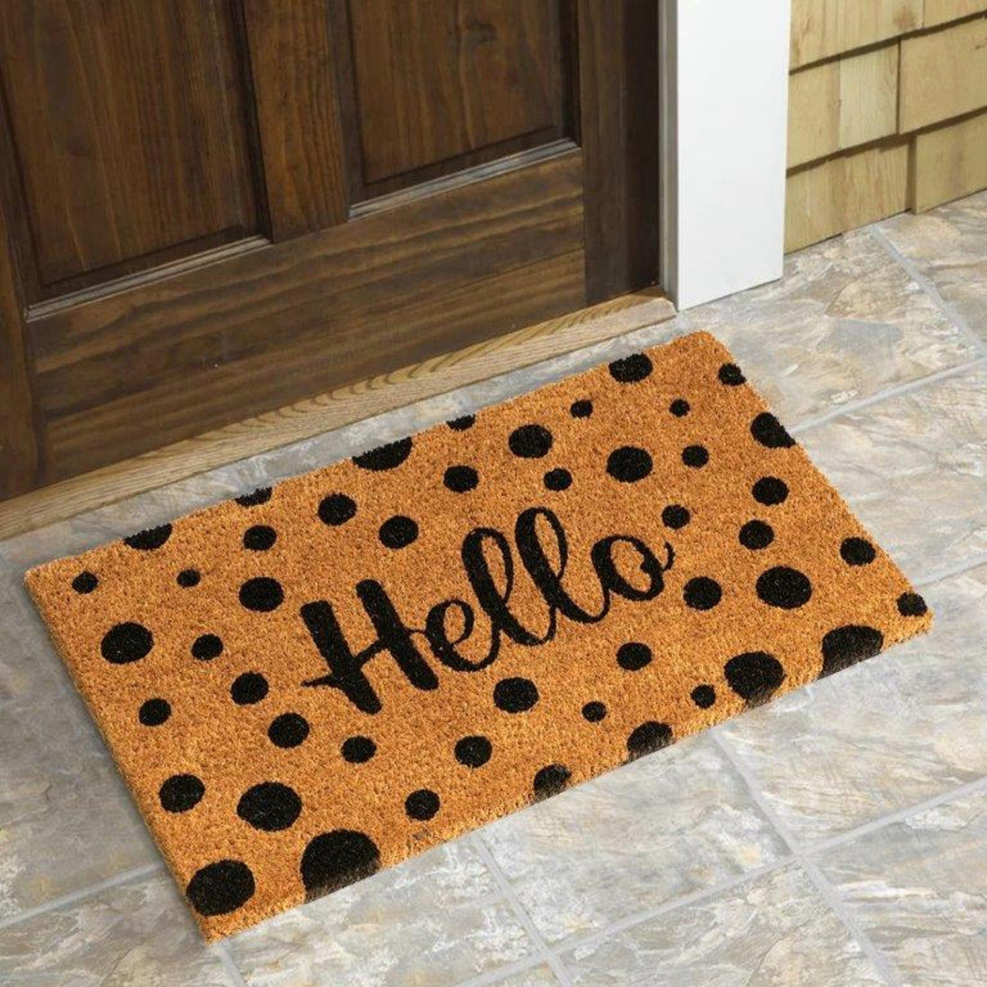 A rectangular natural coir door mat with black polka dots and the word 'Hello' printed on it, placed on a tiled floor by a wooden door.