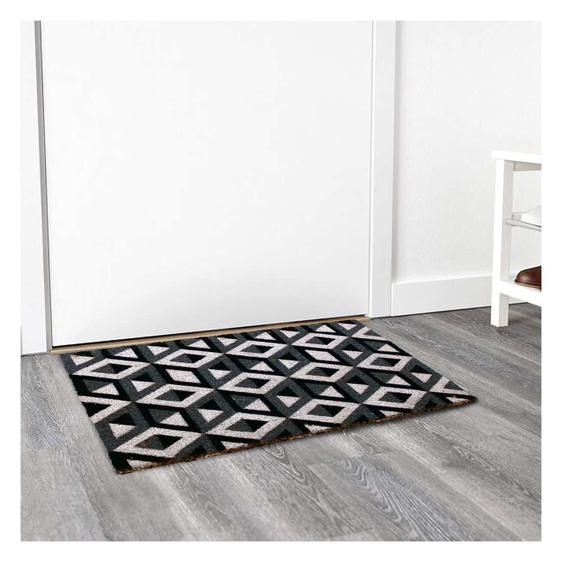 A rectangular natural coir floor mat with a black and white geometric pattern, placed on a floor entrance with a white door in the background.