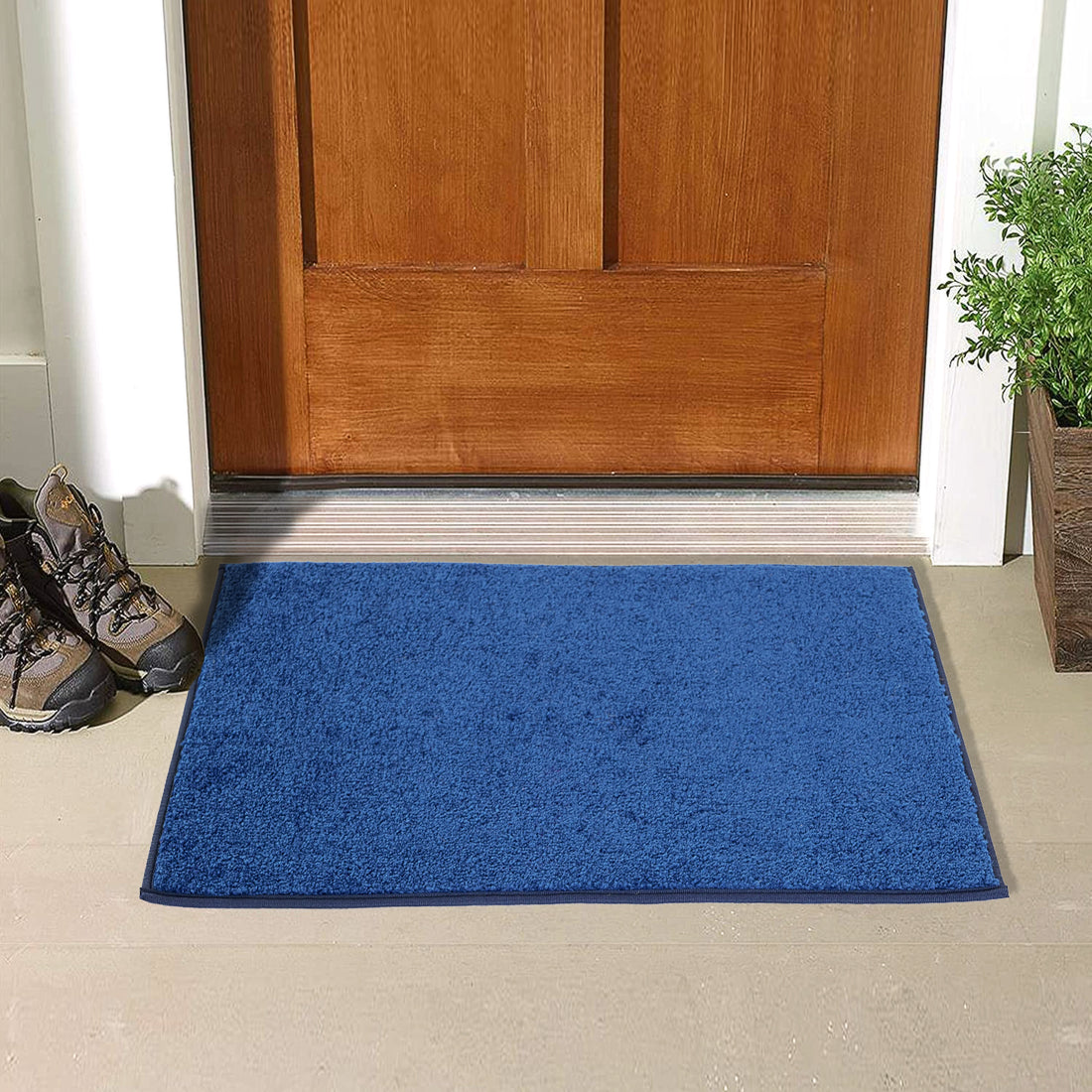 A blue rectangular doormat with a solid pattern is placed on the floor in front of a wooden door, with a pair of boots at the top of the image and a potted plant to the side.