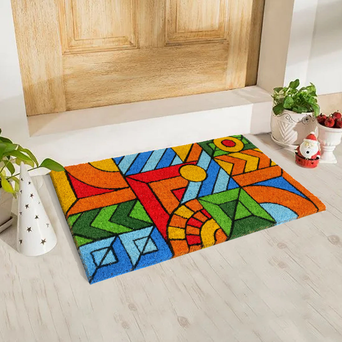 A rectangular natural coir door mat with a colourful geometric pattern, placed on a floor with a wooden door in the background.