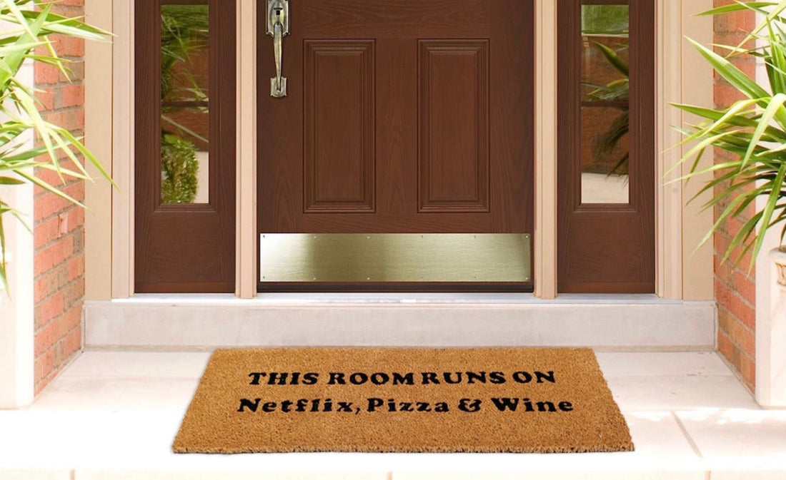 Doormat with text 'THIS ROOM RUNS ON Netflix, Pizza & Wine' in front of a house entrance.