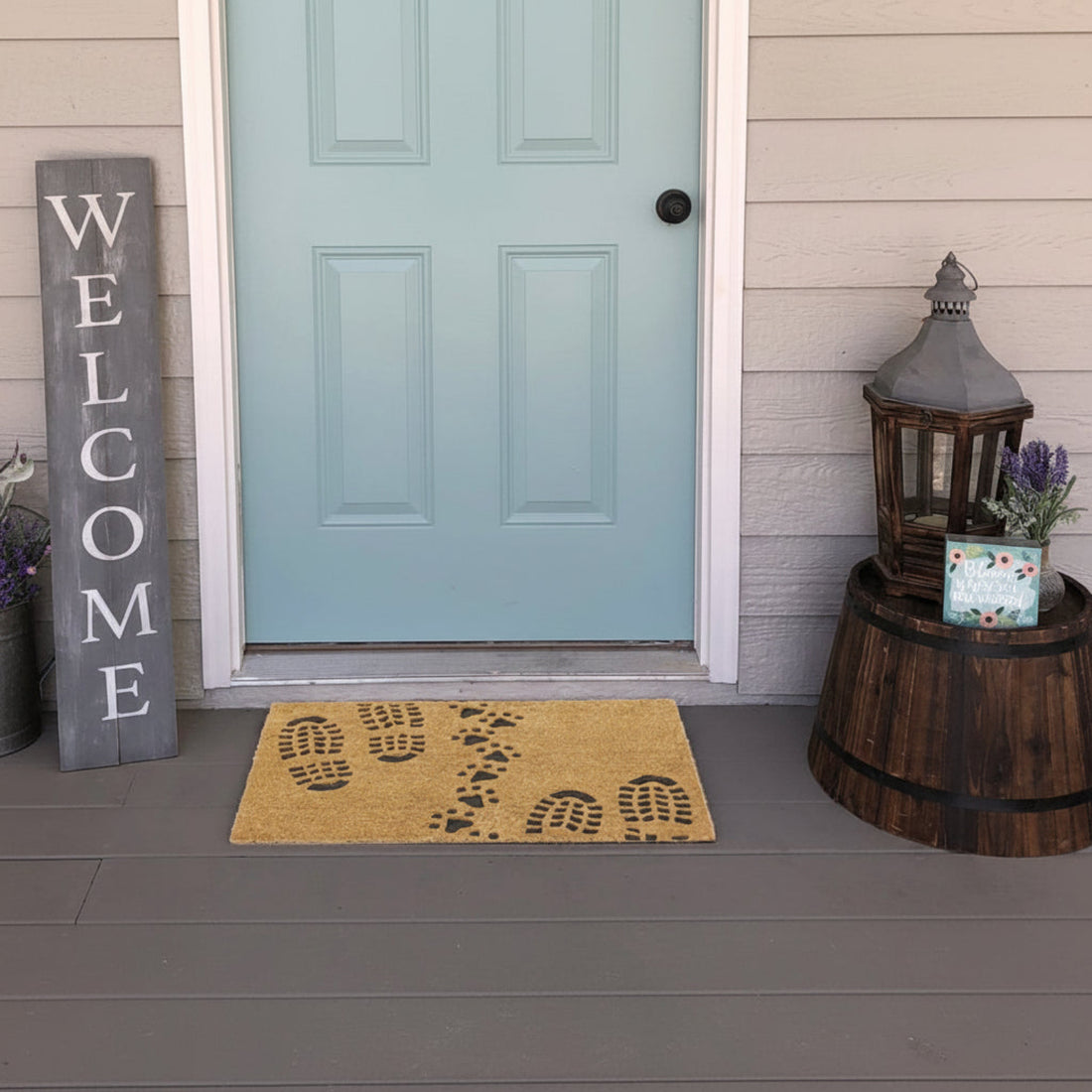 A rectangular coir doormat with a printed design featuring footprints and the word 'WELCOME' on it, placed on the ground in front of a blue door, with a wooden object and a purple flower basket to the side.