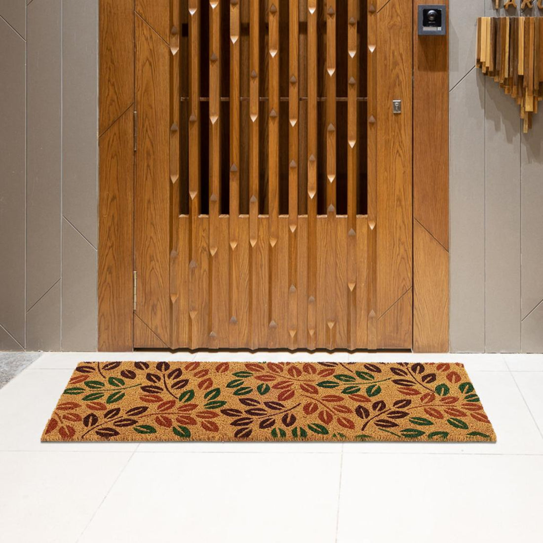 A rectangular natural coir entrance door mat with a leaf printed design, placed on a floor with wooden door panels in the background.
