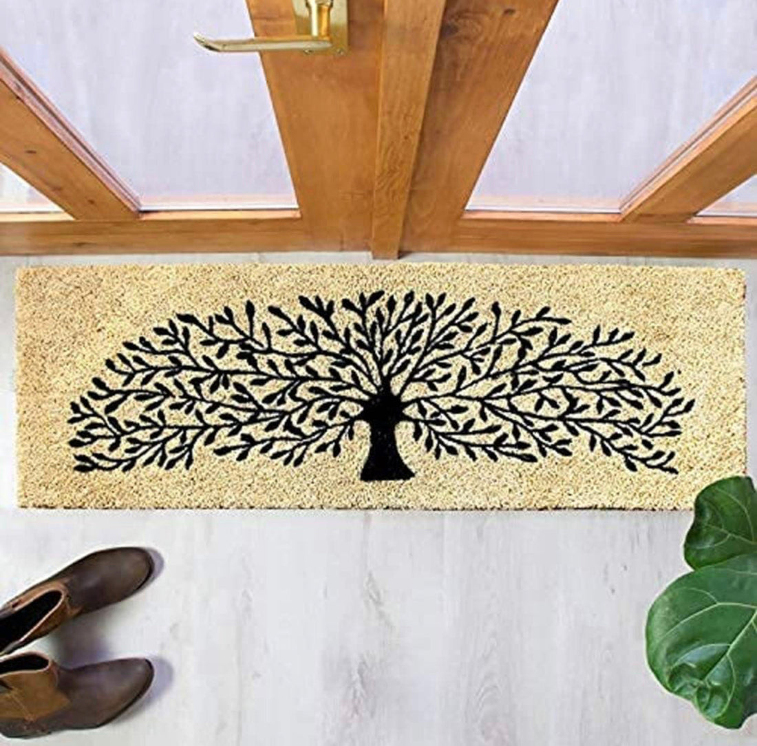 A rectangular, black-colored coir floor mat with a printed design of a tree, placed on the floor by a doorstep, with a pair of shoes at the bottom of the image.