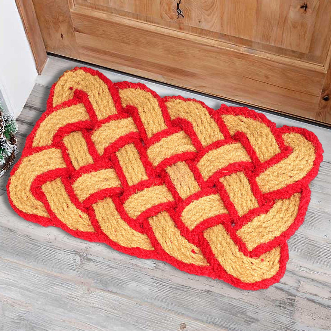 A traditional red and yellow handloom coir rope mat with a knotted geometric pattern, placed on a wooden floor next to a door.