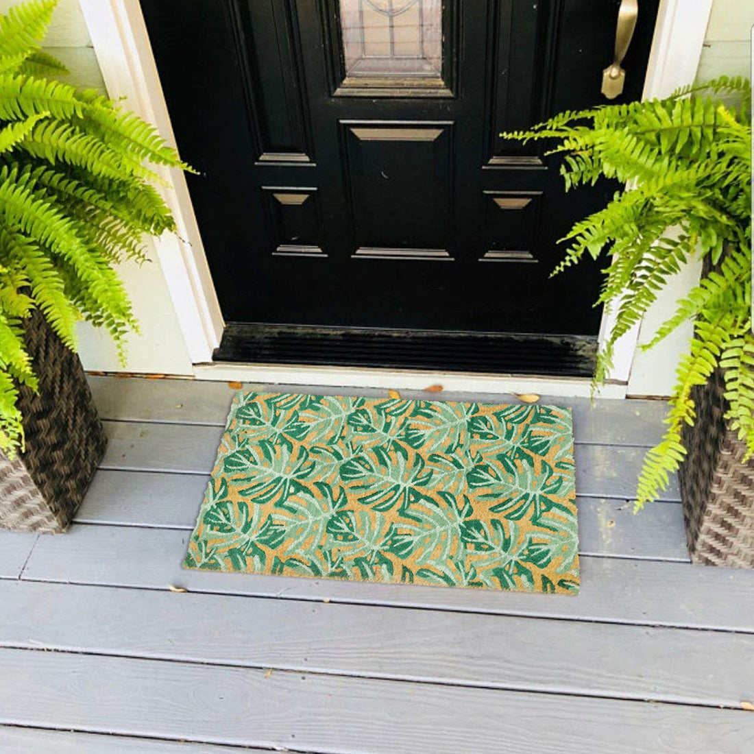 
                      
                        A green coir floor mat with a fern leaf design, placed on a doorstep with fern plants on either side.
                      
                    