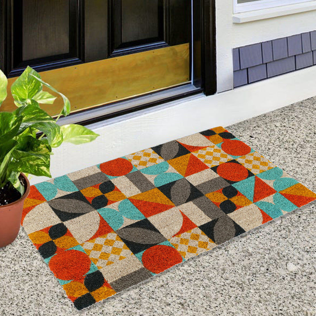 A rectangular door mat with a colourful mosaic pattern featuring orange, yellow, and other pastel colours, placed on a doorstep with a potted plant to the left and a black door in the background.
