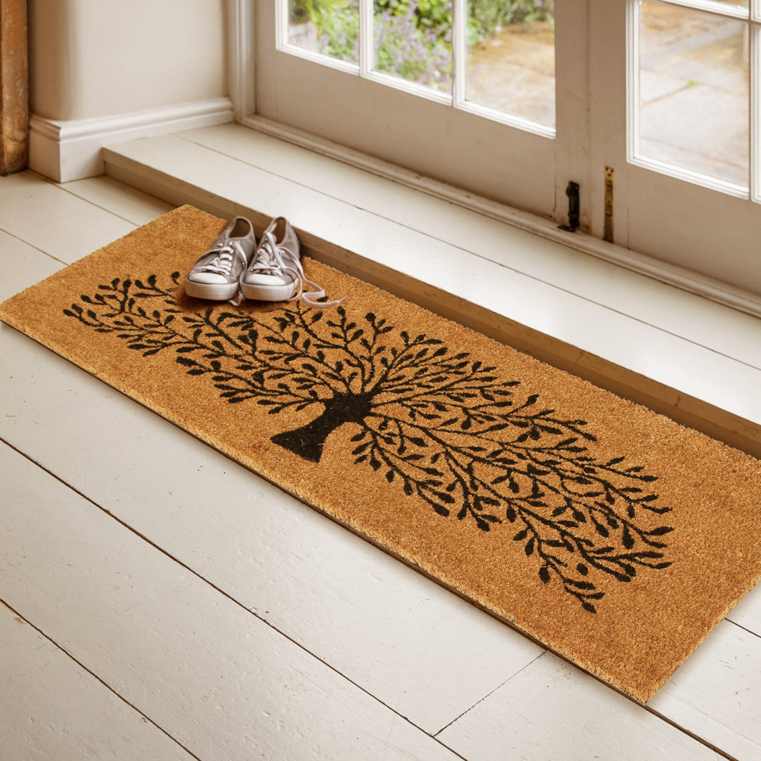 A beige OnlyMat Black Tree Coir Doormat with an elegant tree design placed on a floor, partially under a white door, with a pair of shoes on top of it.
