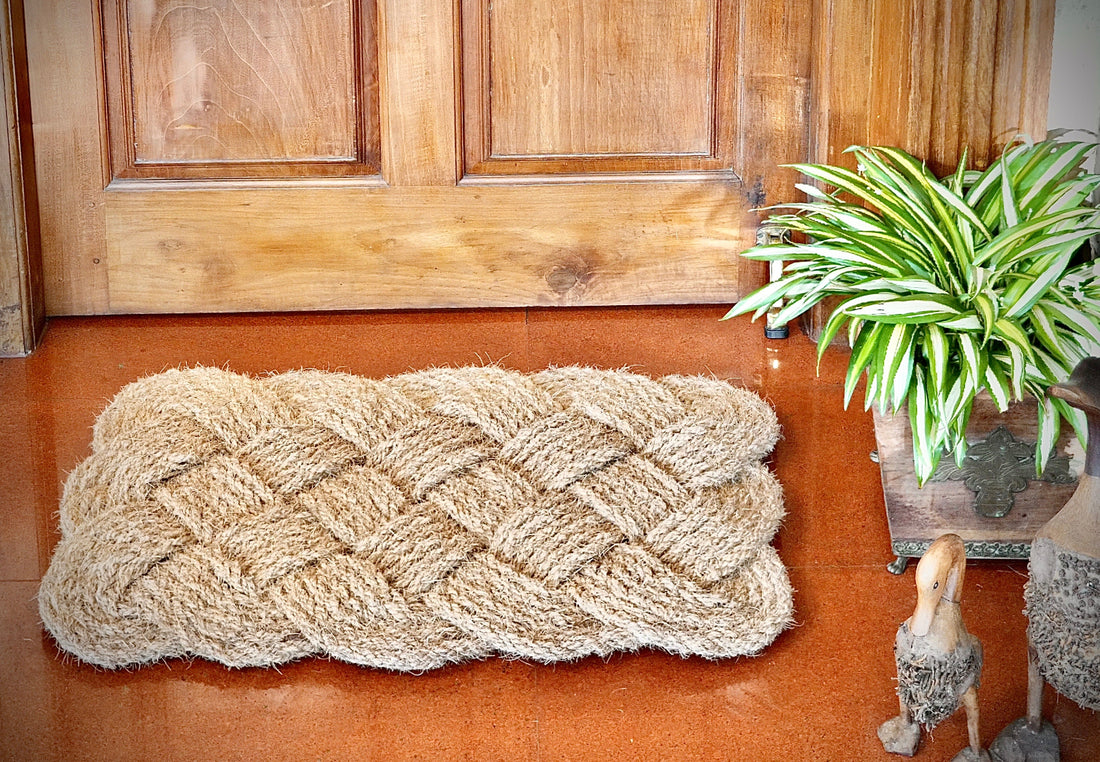 
                      
                        Textured coir doormat on a wooden floor with a plant and decorative items in the background
                      
                    