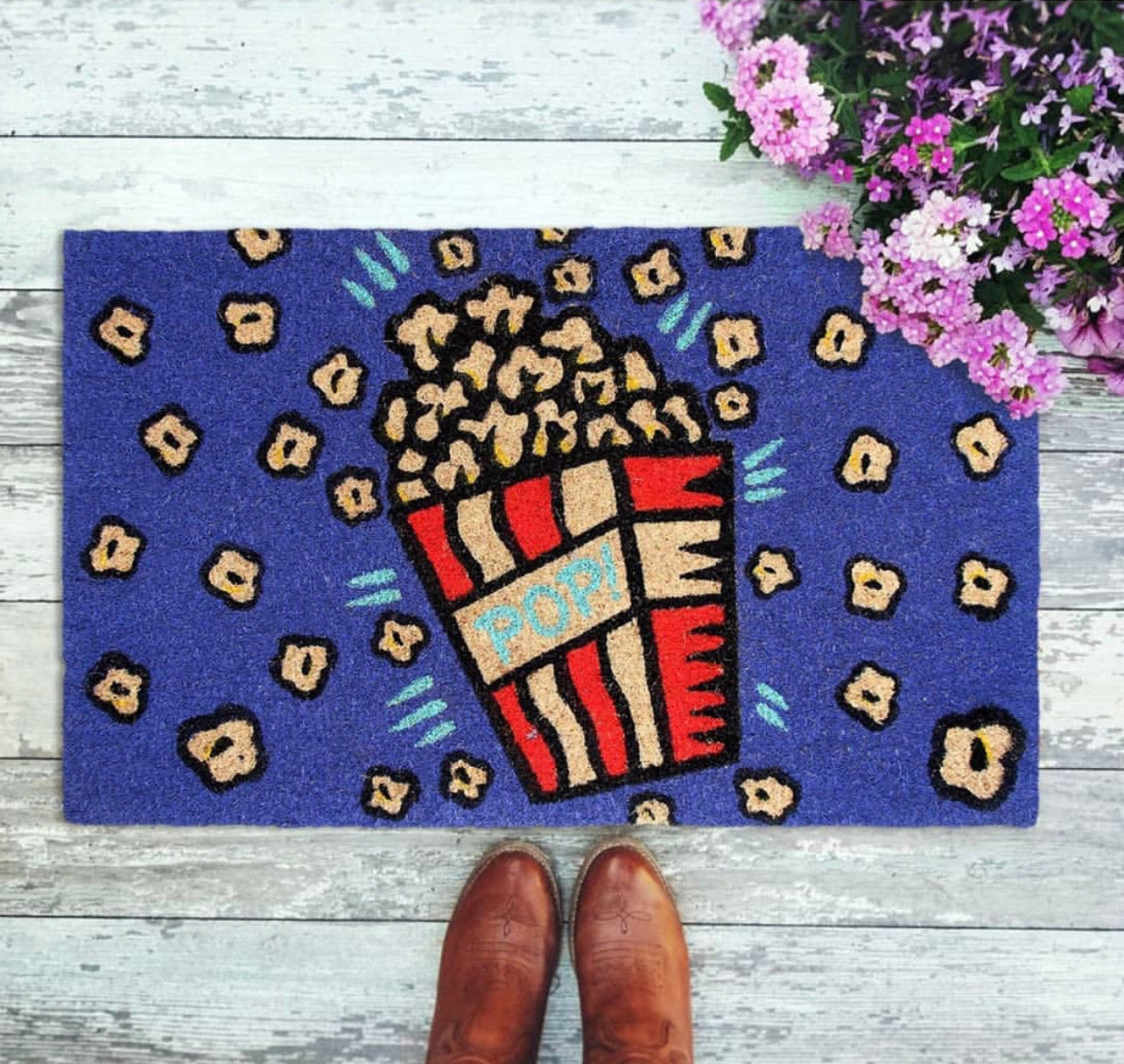 Rectangular natural coir door mat with a colorful popcorn design, placed on a wooden floor, with a person's boot print on the mat.