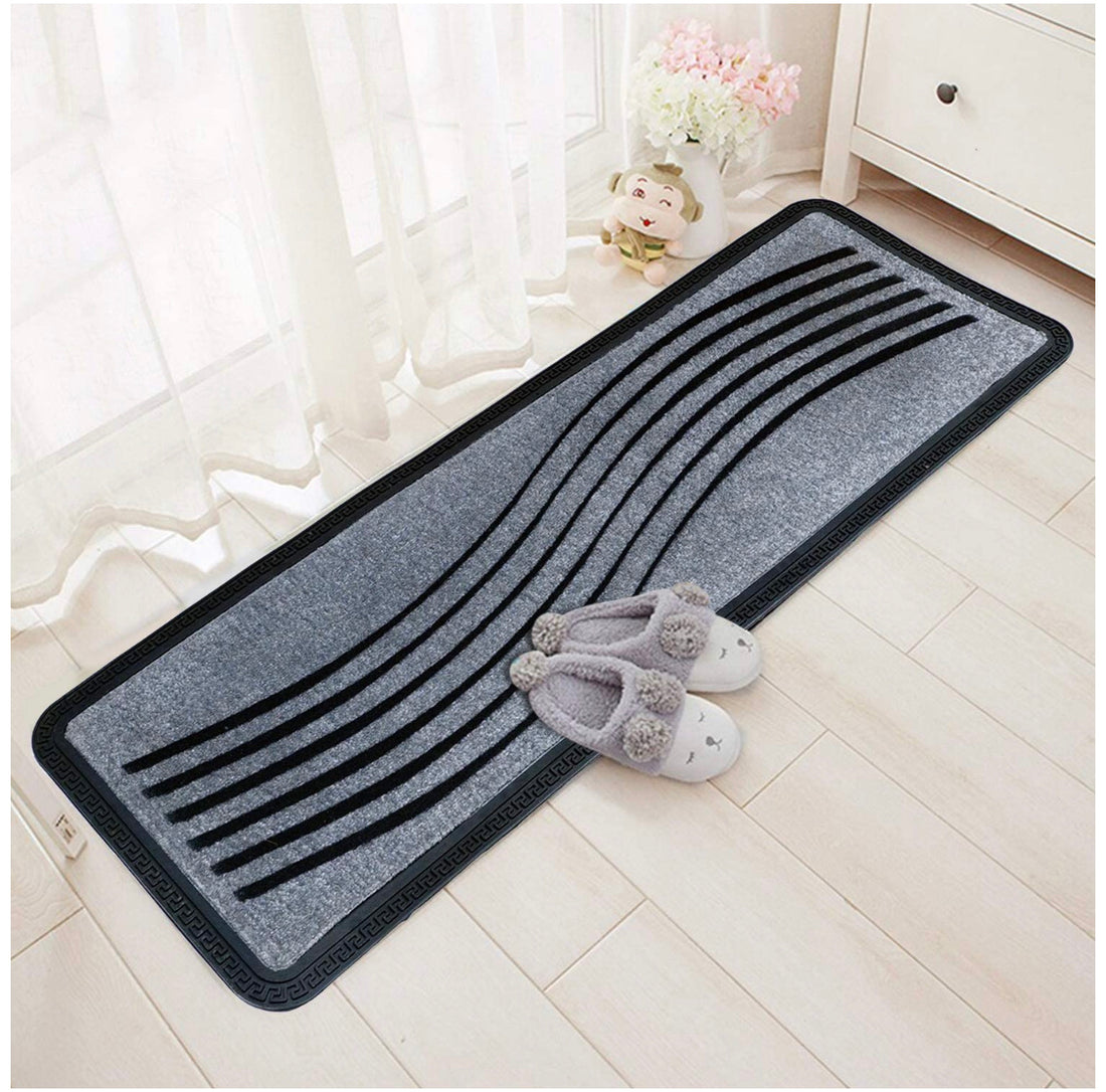 A rectangular, oblong shaped doormat with a silver color and black wave-like pattern, placed on a wooden floor, with a pair of slippers at its bottom corner.